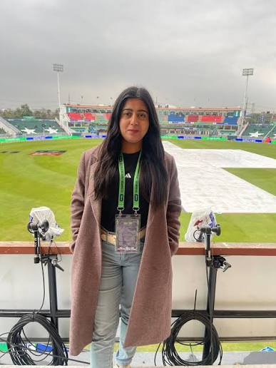 Kiran Khan on location at a stadium, wearing a wool coat and media accreditation